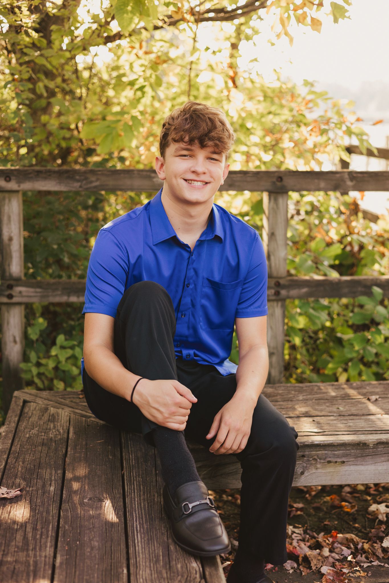 Smiling teenage boy in blue polo shirt sitting on wooden bench outdoors with green foliage background.