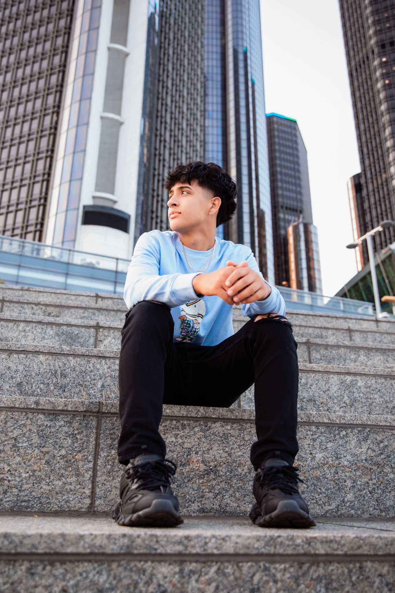 Young man in blue sweatshirt sitting on granite steps with modern skyscrapers in the background.