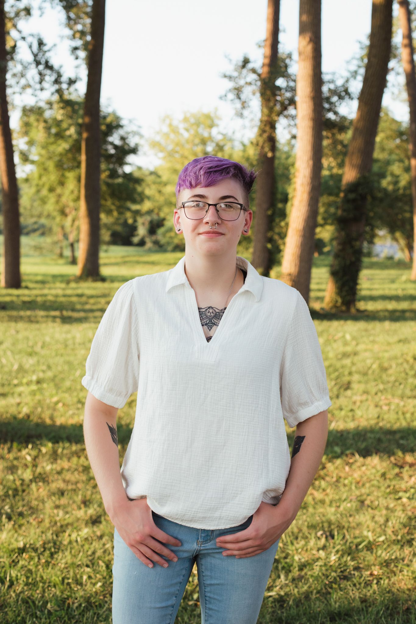 Young person with purple hair and glasses wearing white shirt standing in sunny park with tall trees.