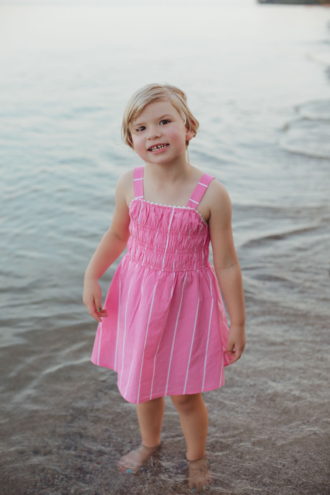 Young girl in pink striped sundress standing barefoot on rocky shore with ocean waves in background.