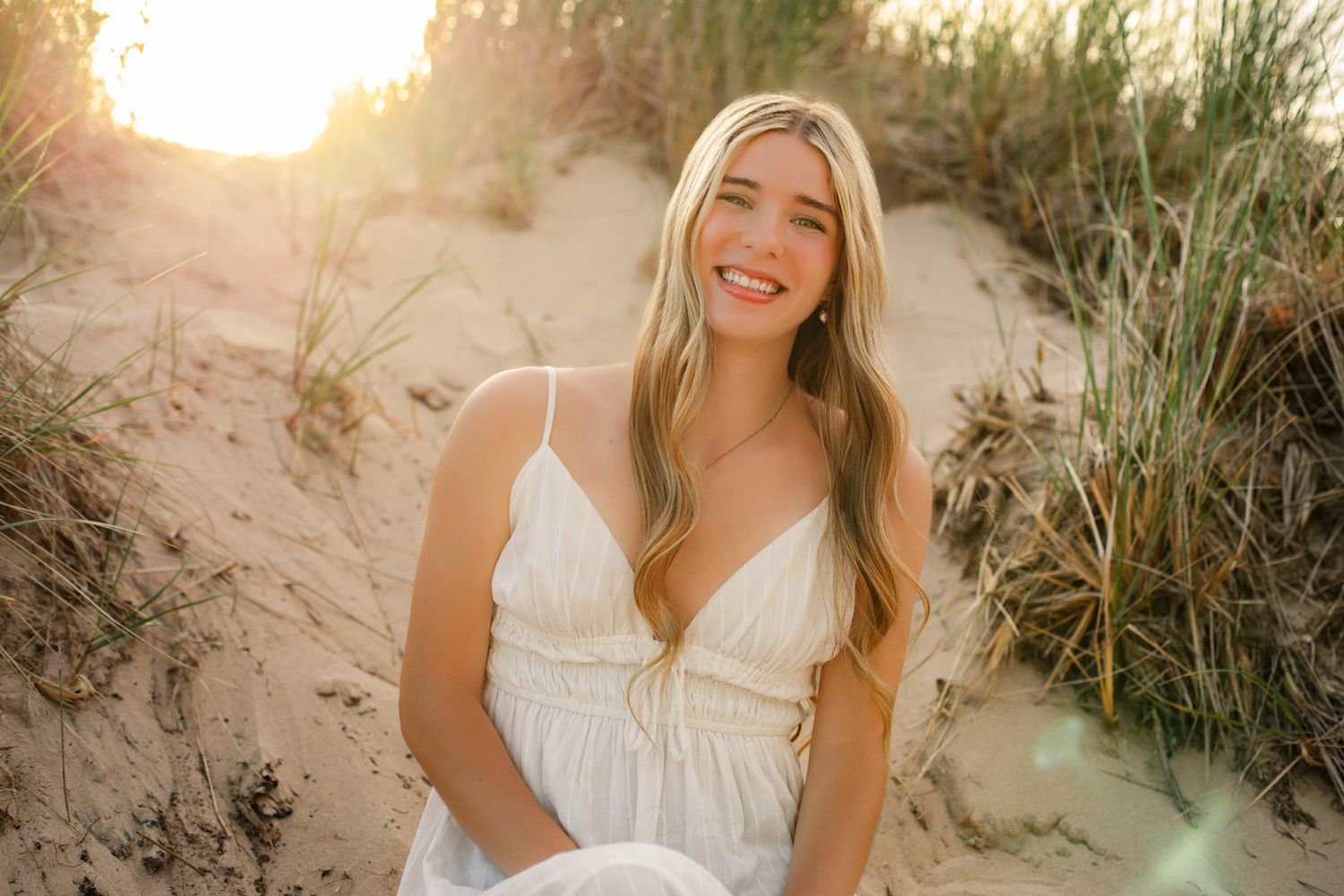 Cheerful blonde woman in white dress sitting on sand dunes with golden sunset light and beach grass.