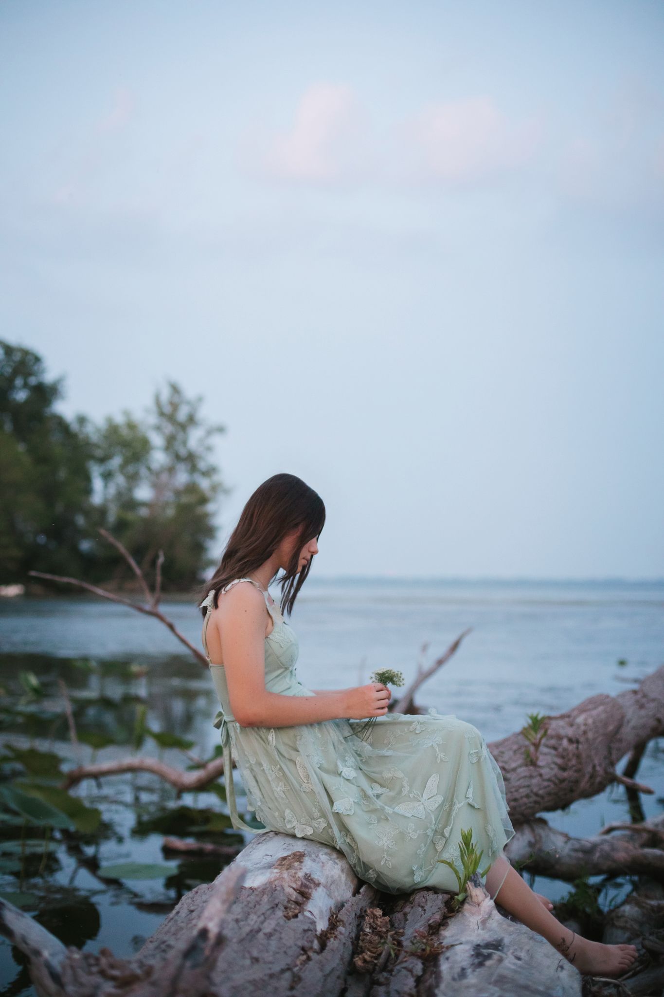 Young woman in mint dress sitting on driftwood by calm water at dusk, looking down.