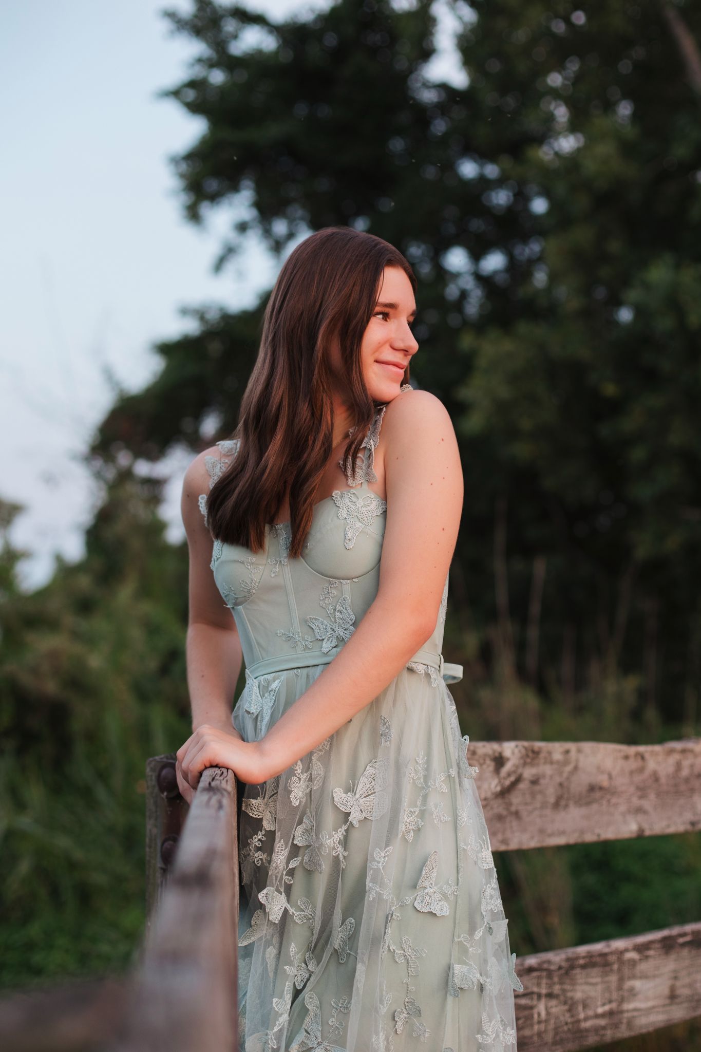 Young woman in mint floral dress leaning on rustic wooden fence outdoors at golden hour.