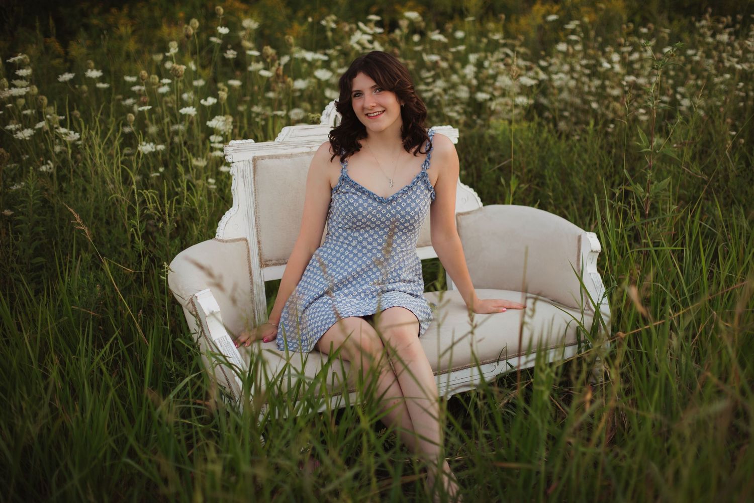 Young brunette woman in blue gingham dress sitting on a white vintage sofa in a wildflower field.