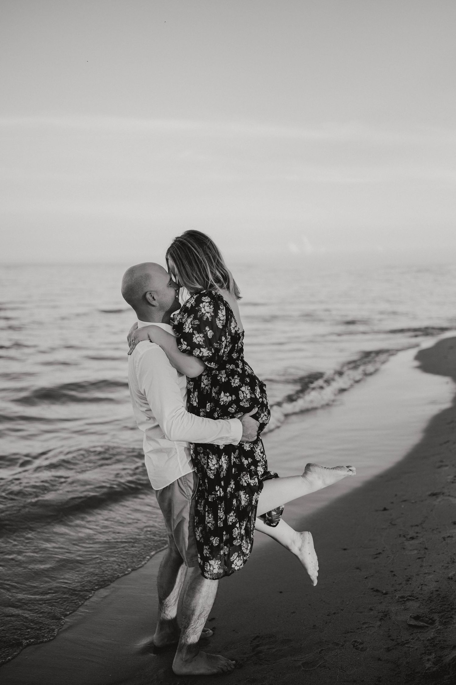 Black and white photo of a man lifting his partner on a sandy beach by the ocean waves.