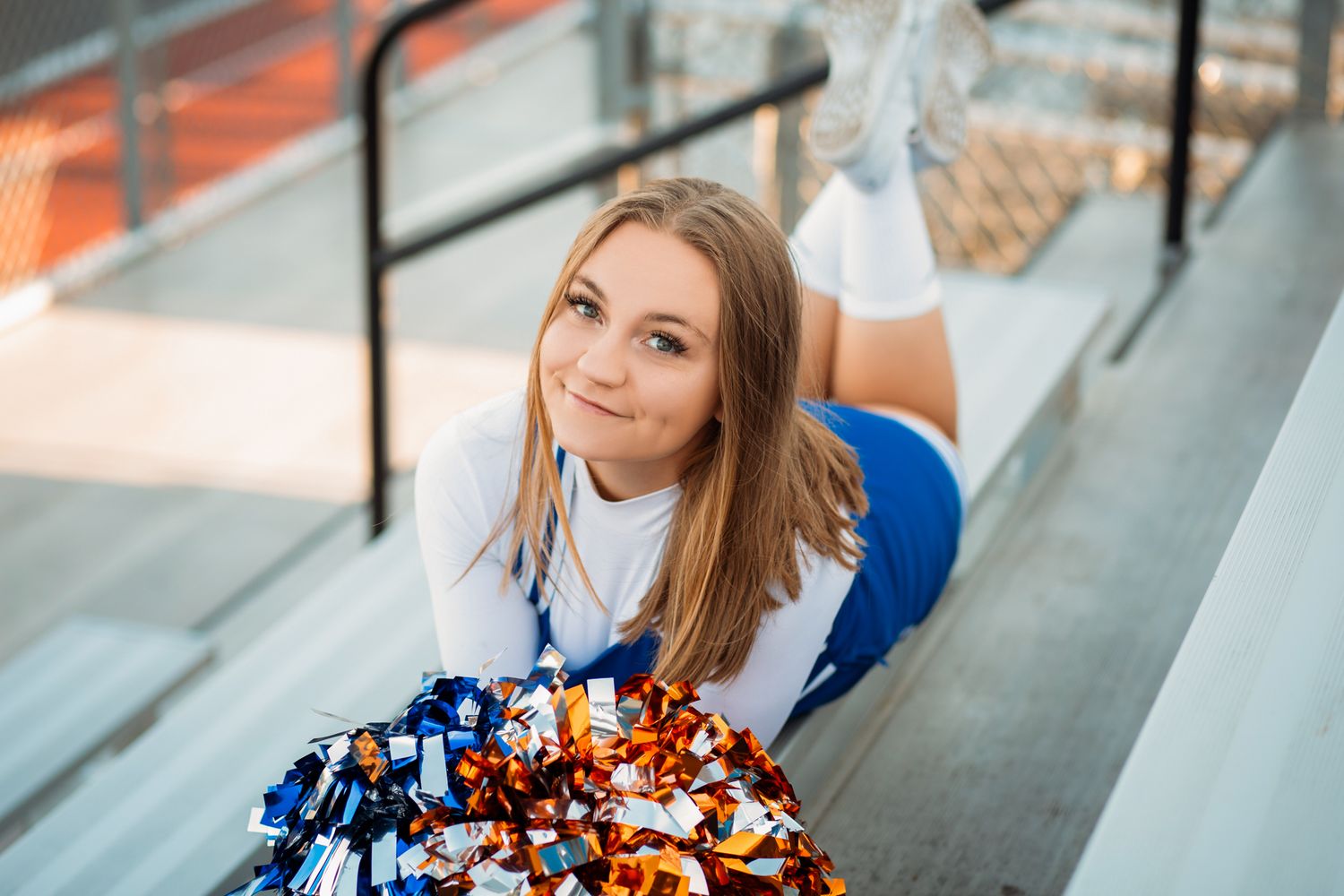 Cheerleader in blue and white uniform poses with colorful pom-poms on stadium bleachers.