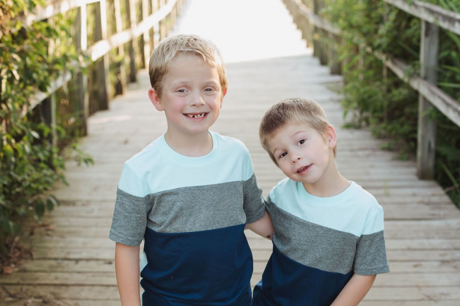 Two smiling boys in matching striped shirts pose together on a wooden boardwalk near the beach.
