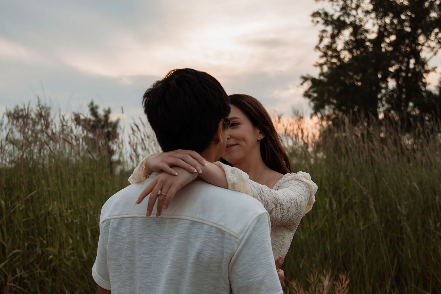 Couple embracing in a field at sunset, woman smiling over man's shoulder, wearing a white lace top.