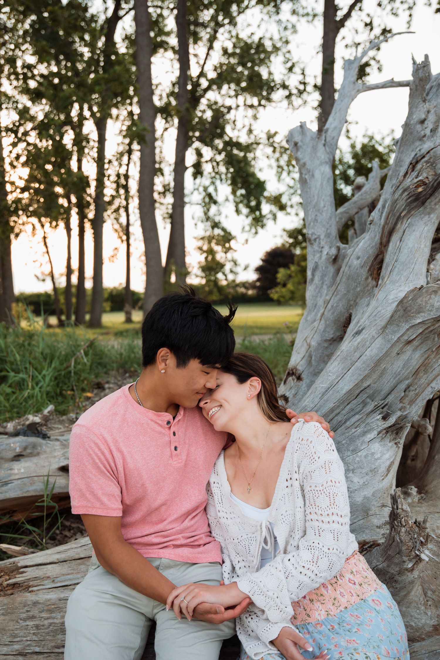 A couple sits together against a driftwood log at sunset, smiling and holding hands in a lush outdoor setting.