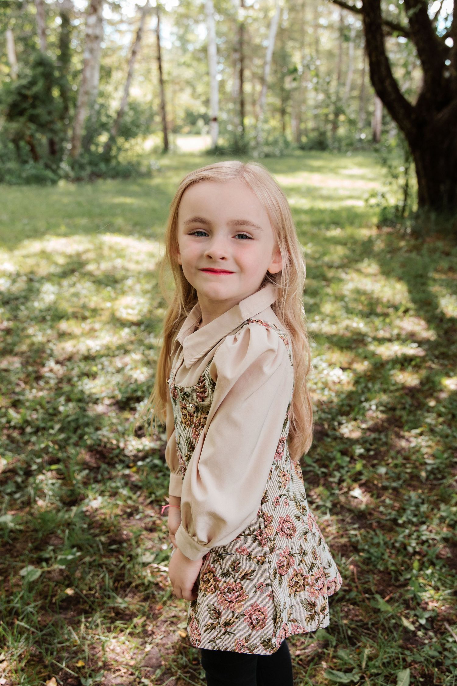 Young blonde girl in floral dress smiling over shoulder in sunny forest setting.