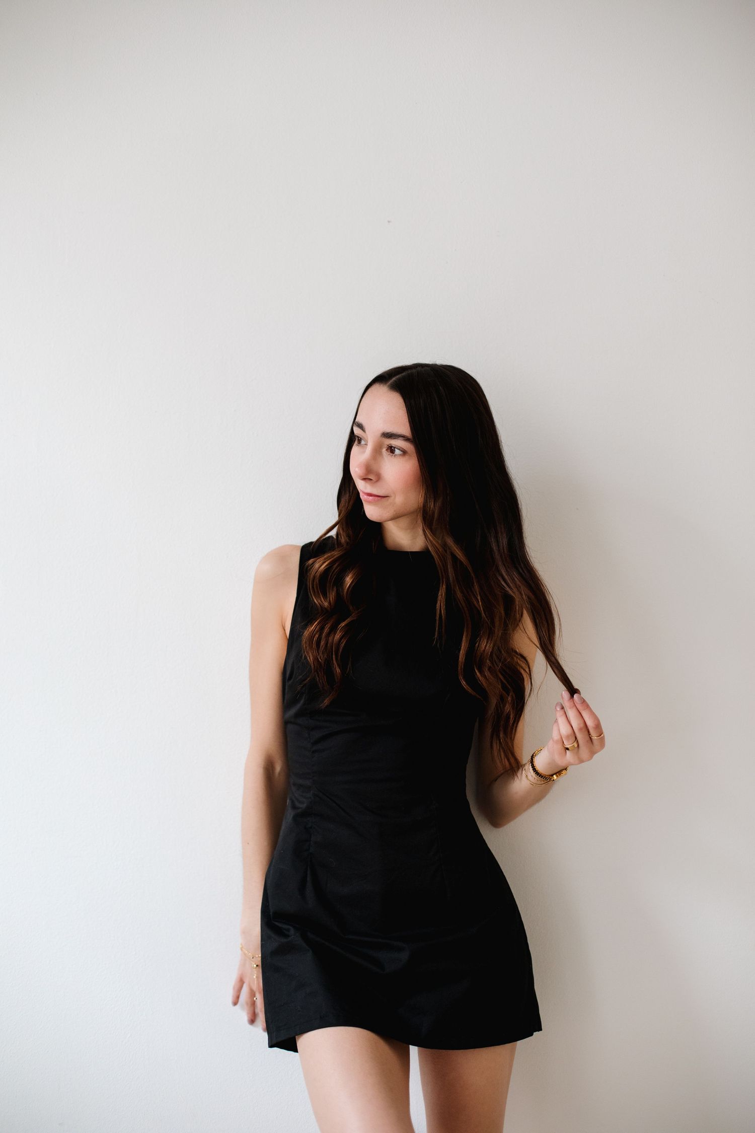 Brunette woman in black mini dress standing against white wall touching her hair.