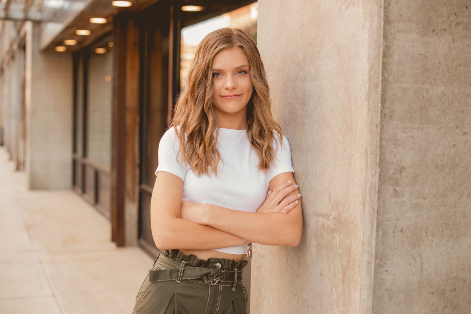 Confident teen girl in white crop top and olive pants leaning against concrete pillar outdoors.