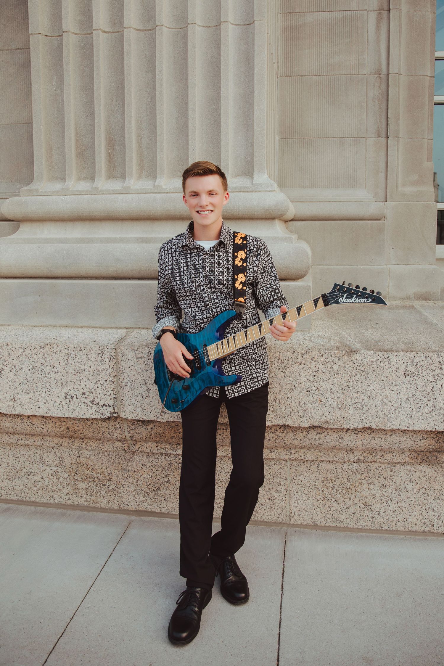 Smiling young man posing with a blue electric guitar and floral strap leaning against stone pillar.