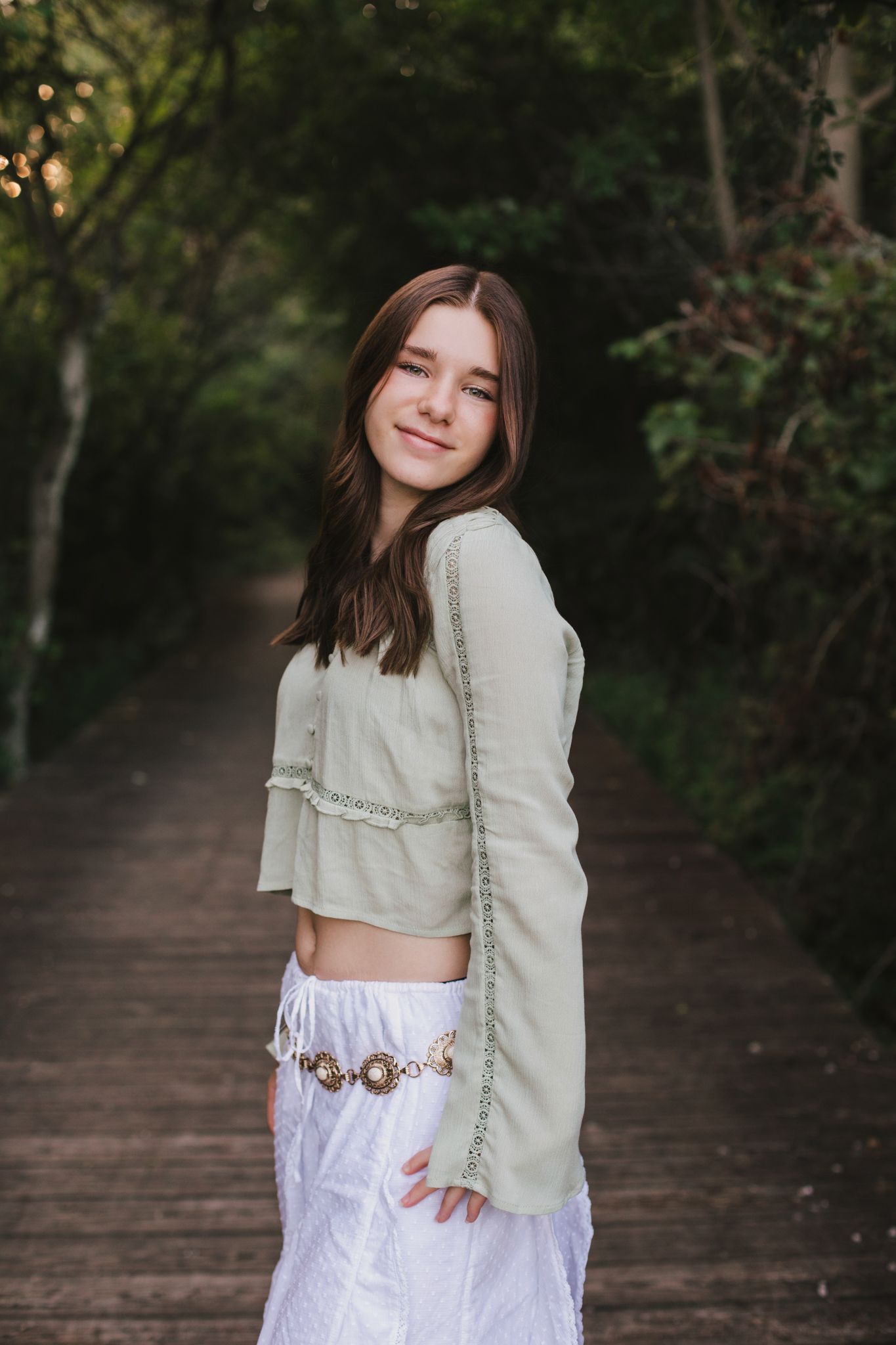 Young girl in sage green crop top and white skirt smiling on a wooden boardwalk surrounded by lush greenery.