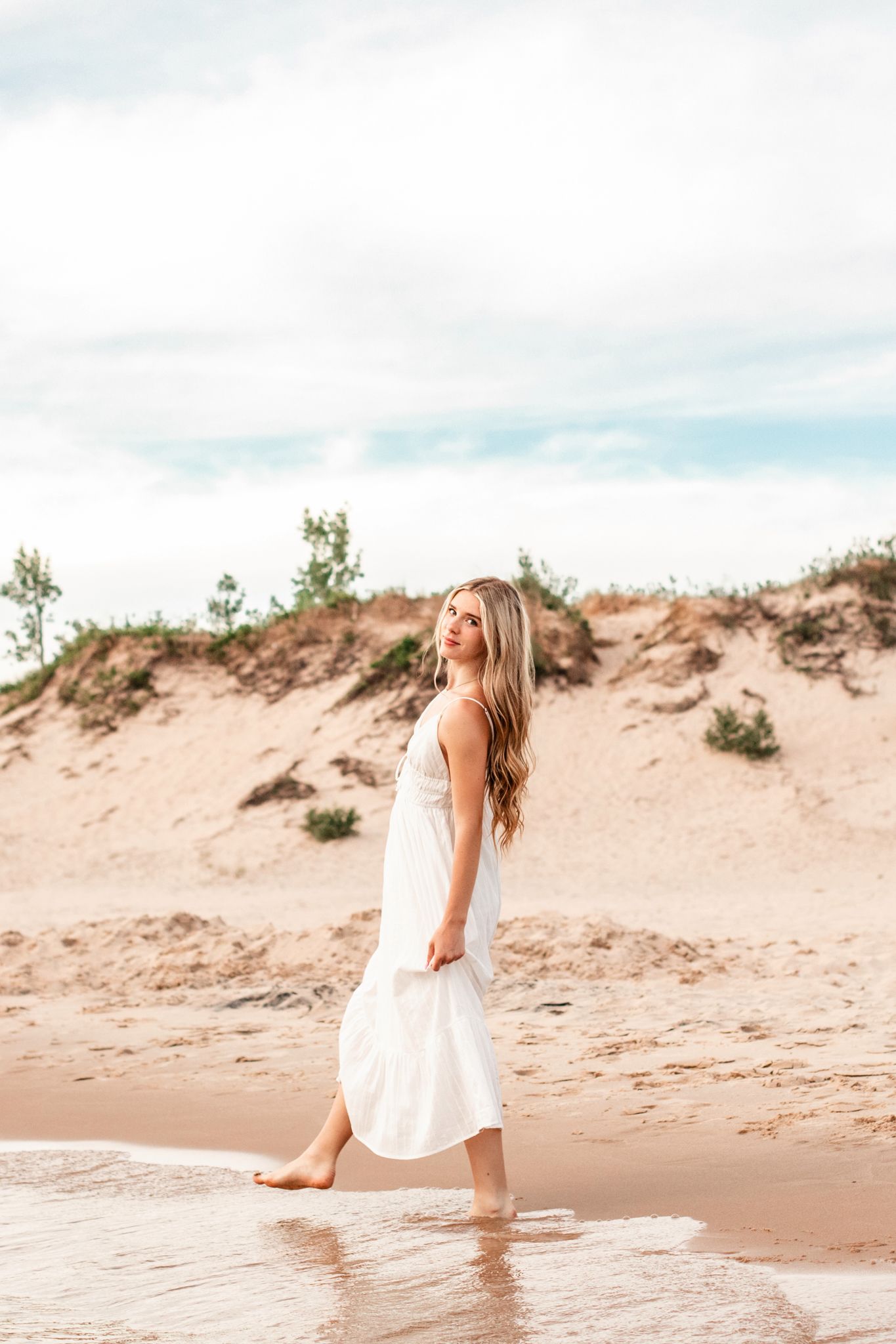 Young blonde woman in white dress walking barefoot on sandy beach shore with sand dunes in background.