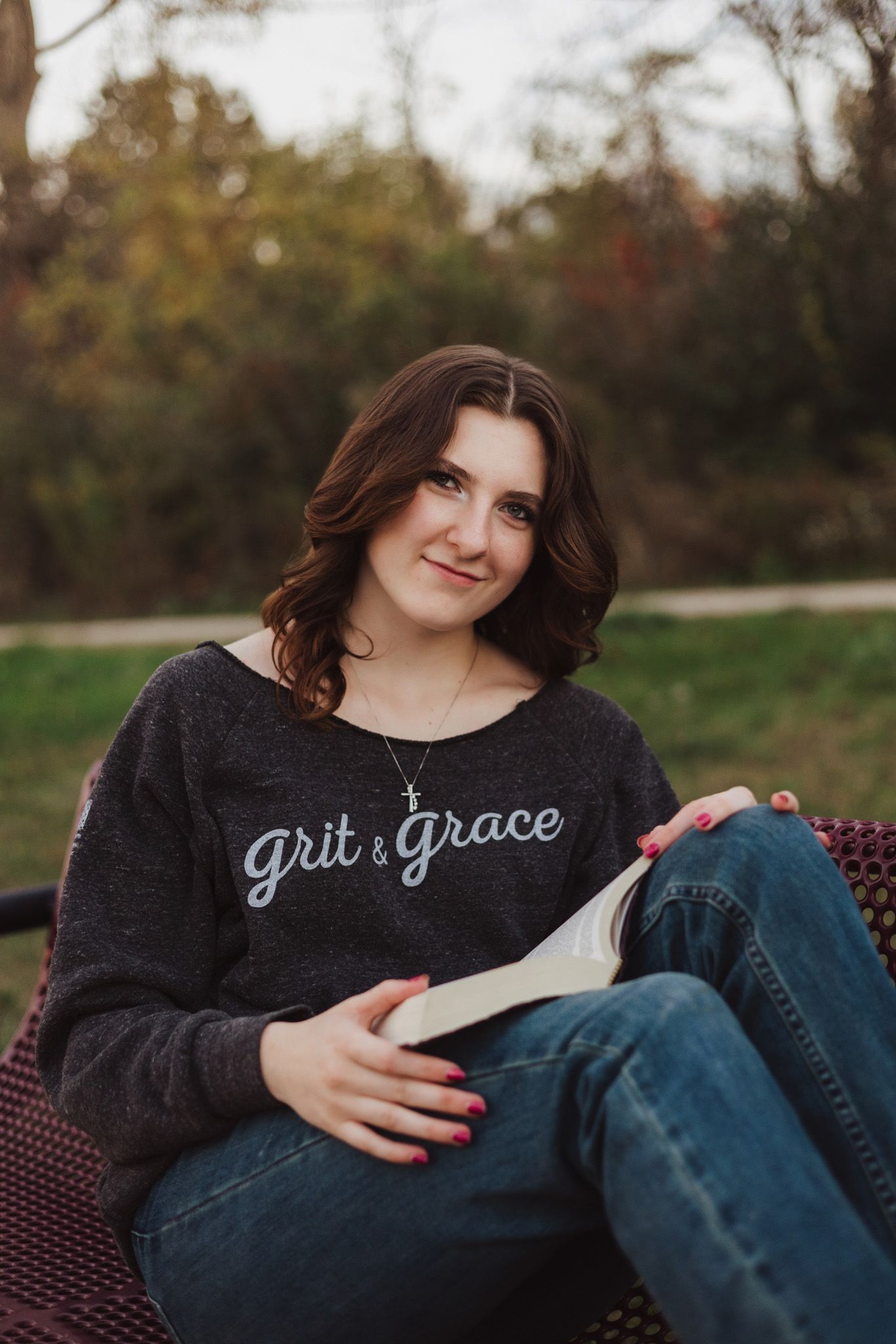 Young woman in a grit and grace sweatshirt reading a book outdoors in a park setting.