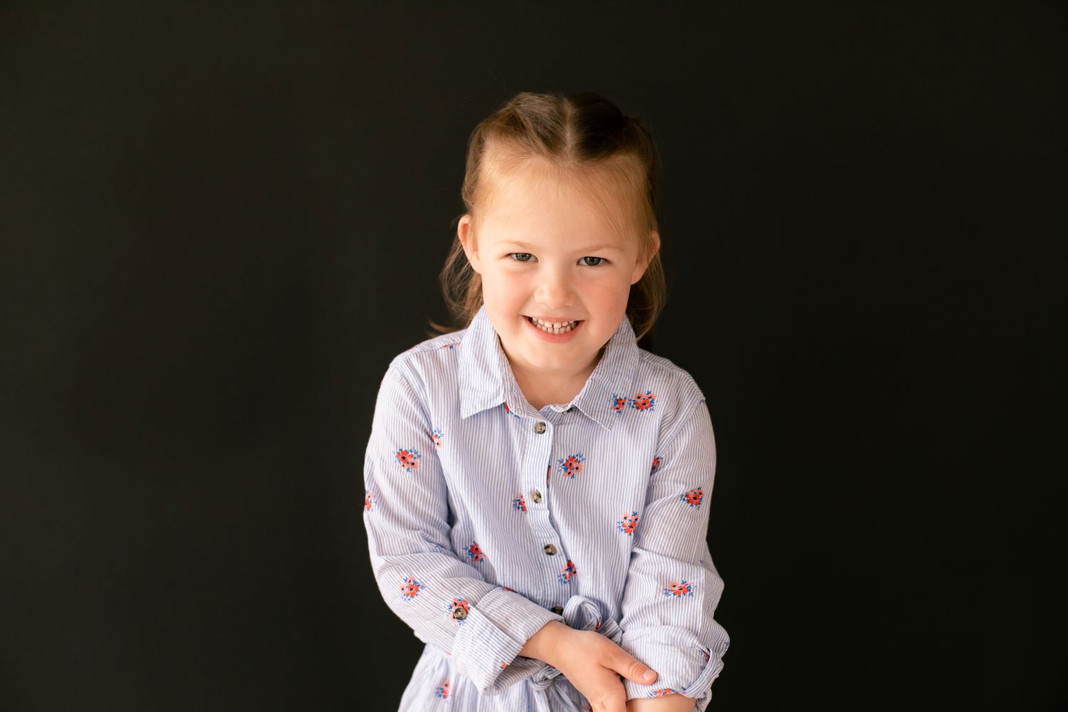 Adorable young girl in floral dress smiling with arms crossed against dark background portrait.