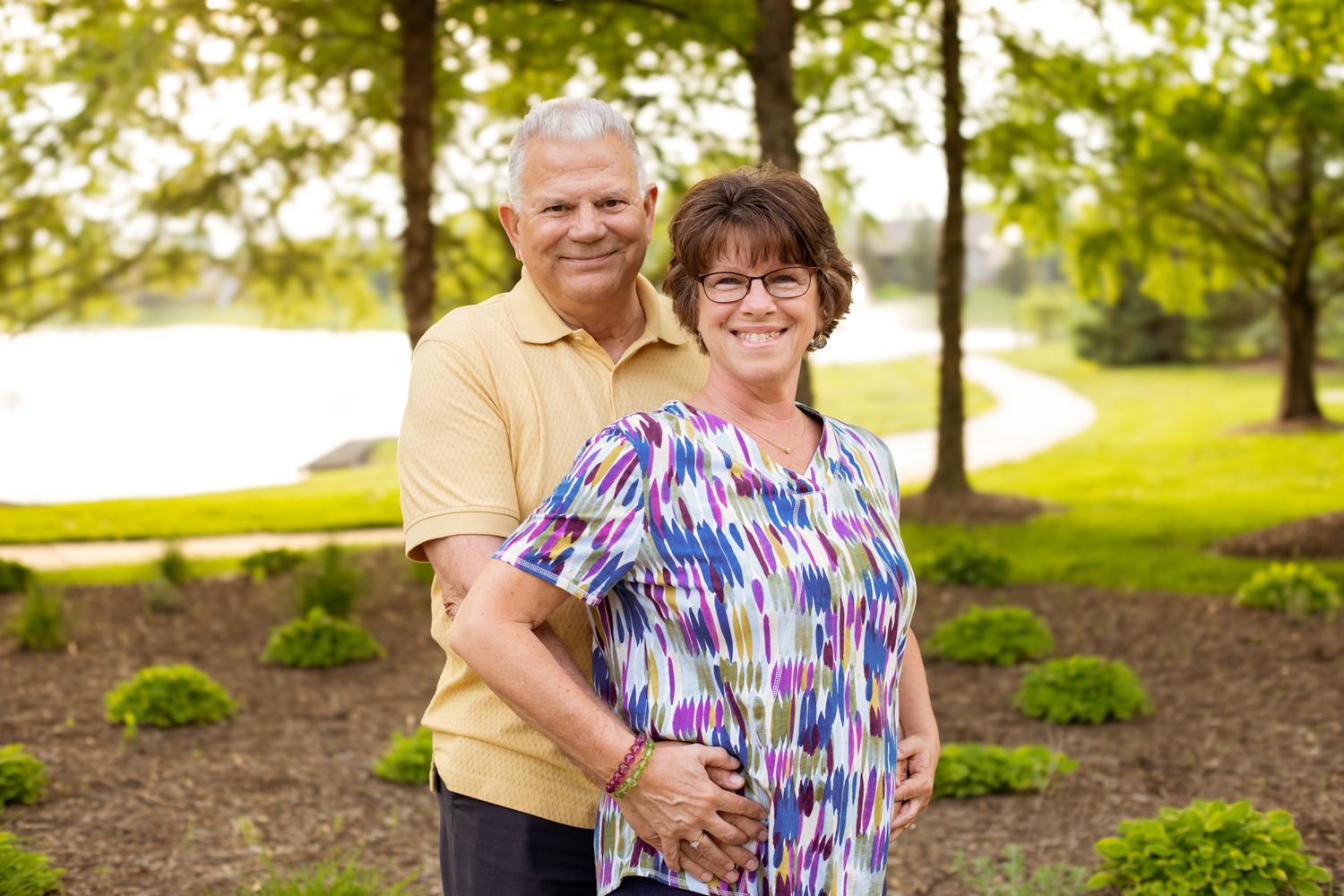 Smiling older couple posing together outdoors in a lush green park setting near a pond.