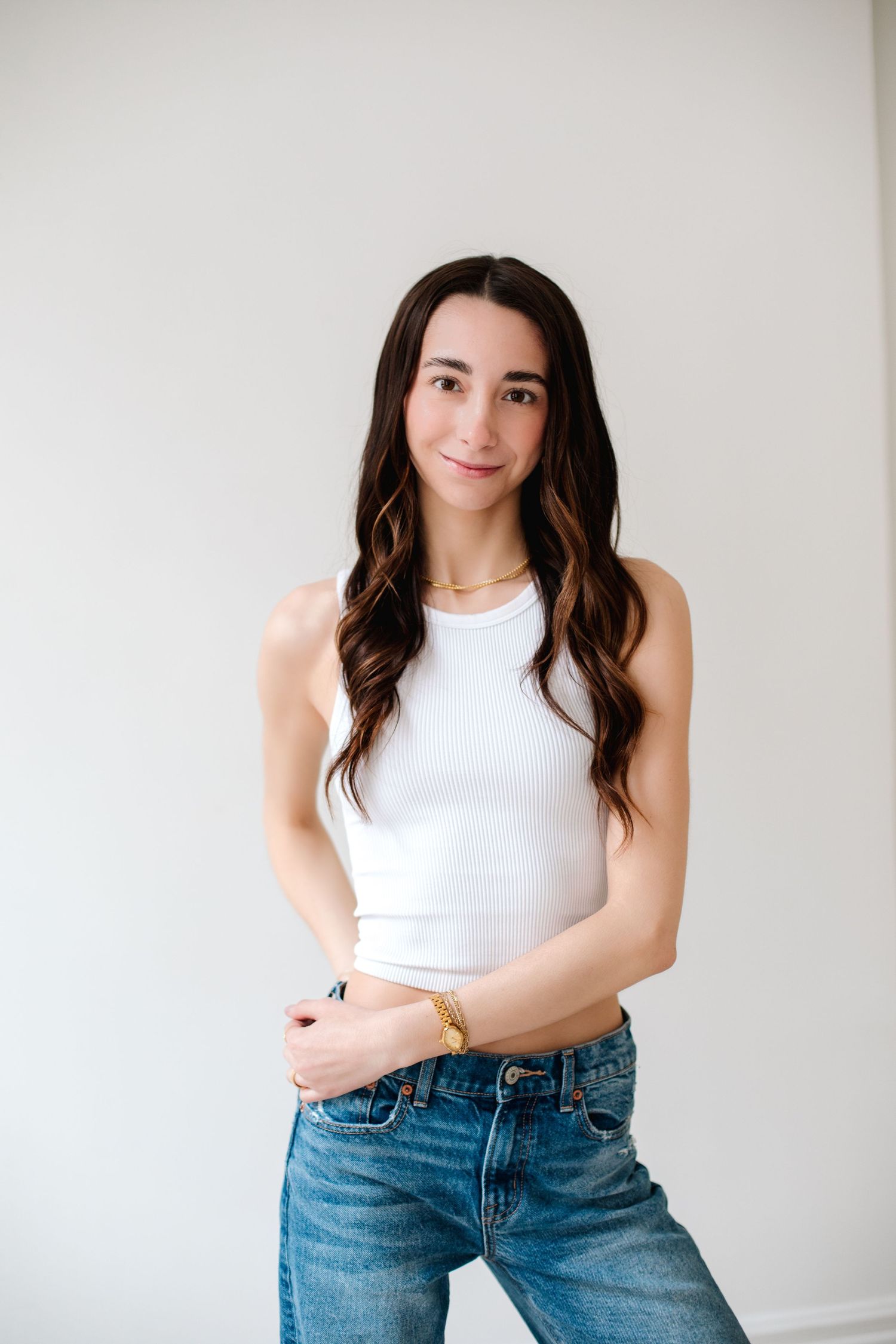 Young woman with long dark wavy hair wearing a white tank top and blue jeans, smiling at camera.