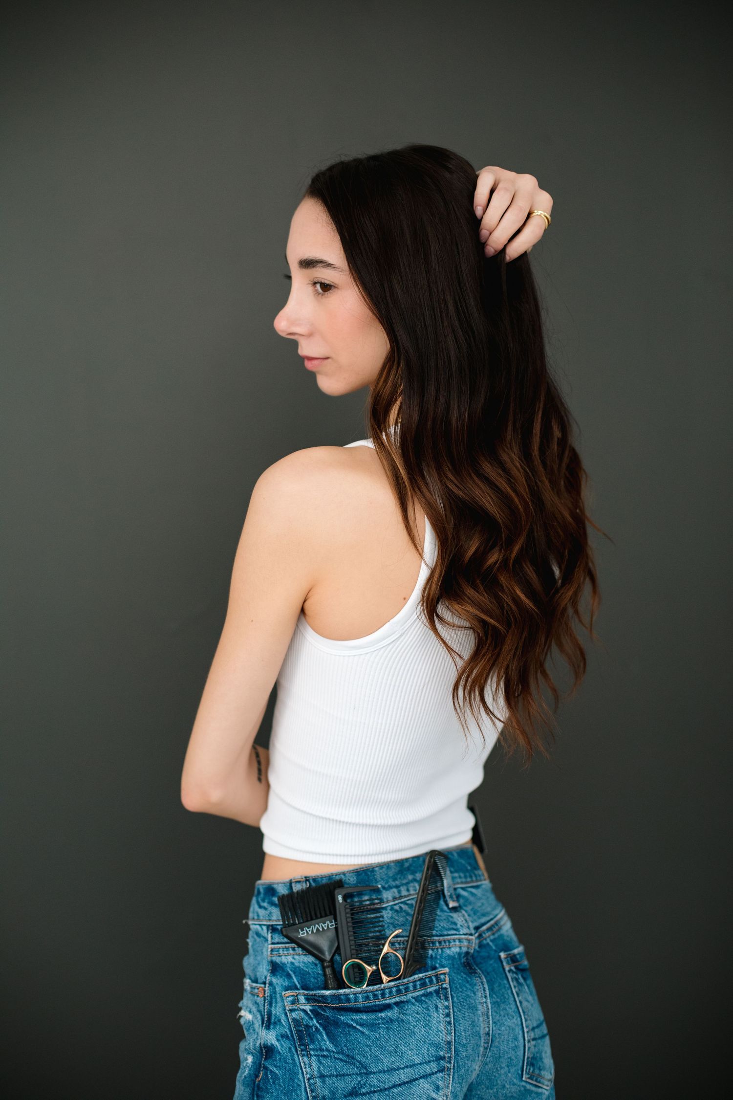 Young woman in white tank top and jeans with hairstyling tools in back pocket, side view.
