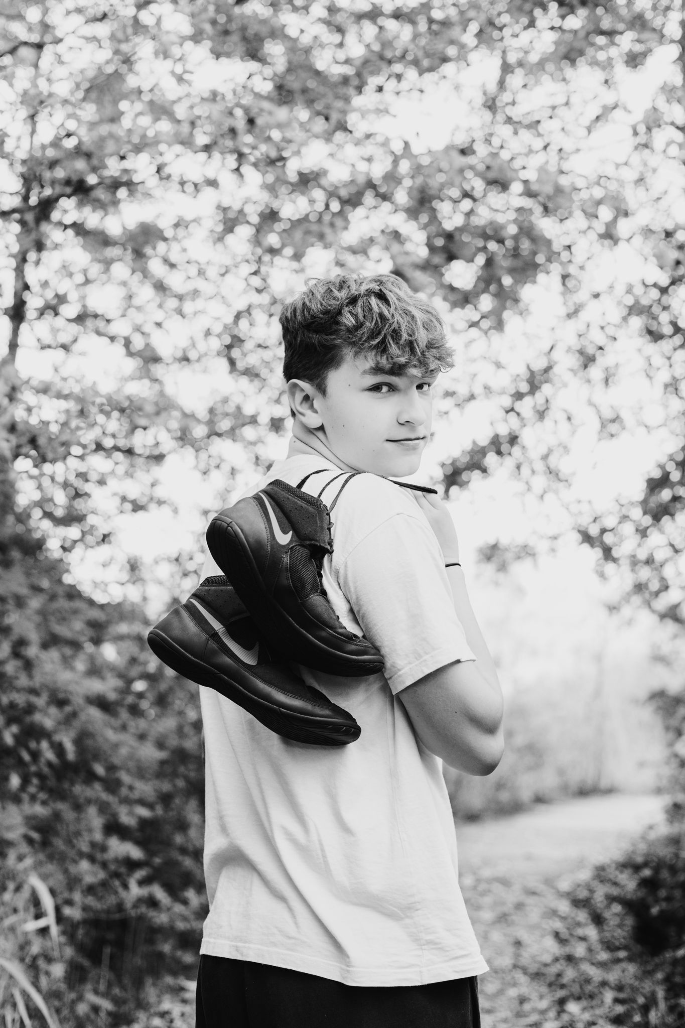 Black and white photo of a teenage boy holding Nike shoes over his shoulder, standing outdoors among trees.