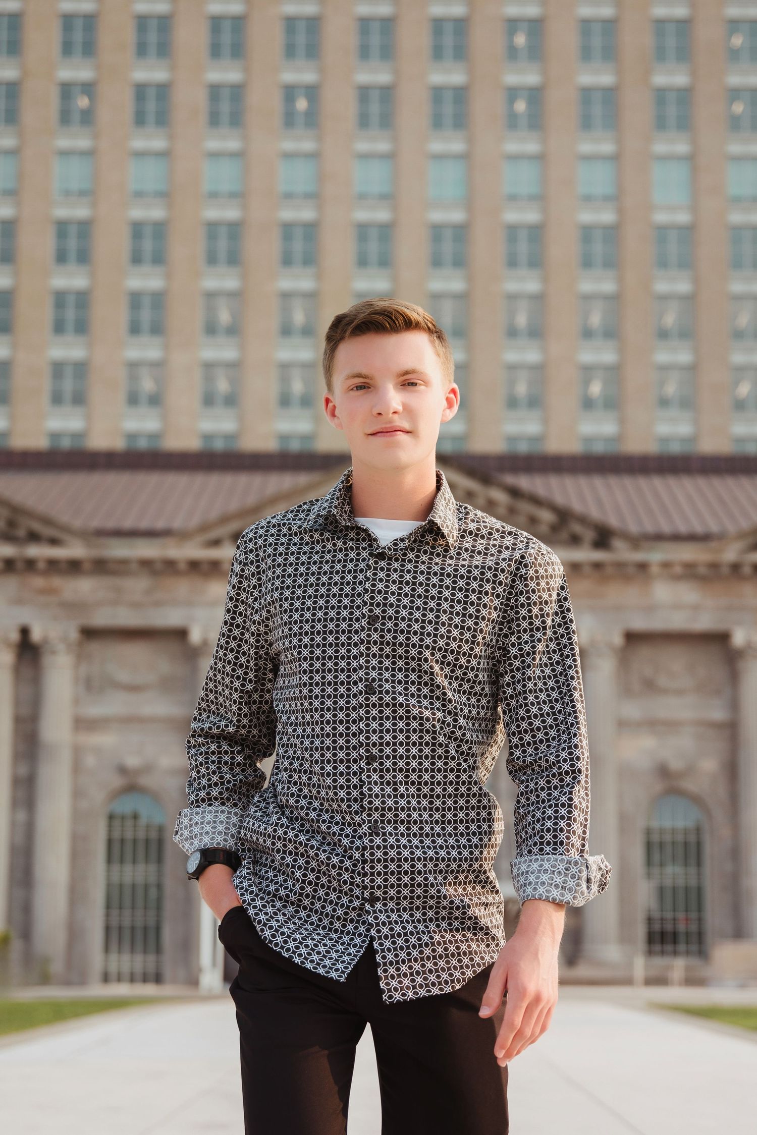 Young man in patterned button-up shirt stands confidently in front of a large historic building.