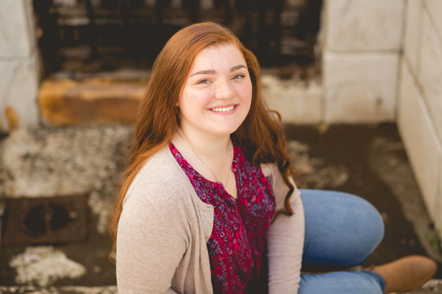 Smiling young woman with red hair wearing a floral top and cardigan, seated outdoors near a stone fireplace.