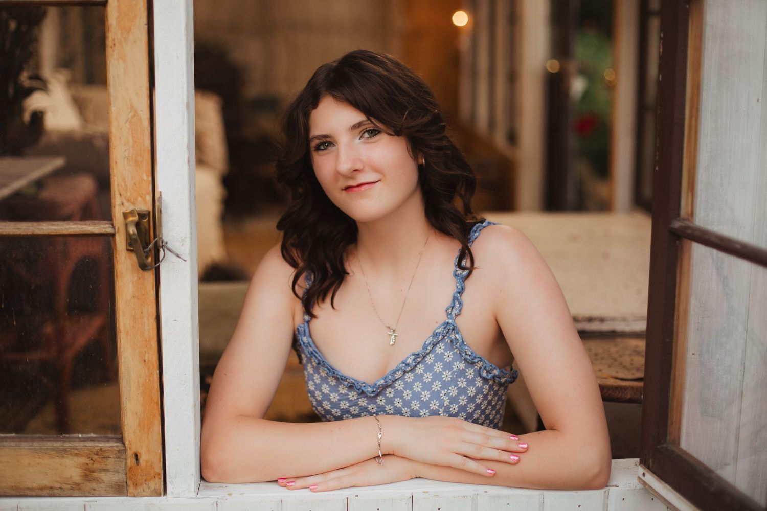 Smiling young woman in blue floral dress leaning on vintage window frame indoors.