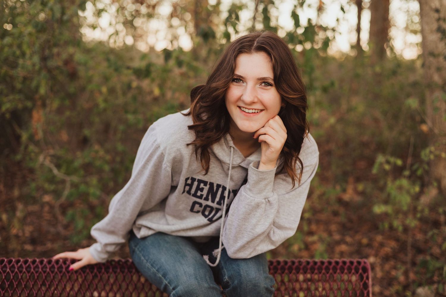 Smiling young woman in Henry College sweatshirt sitting outdoors in autumn woods.