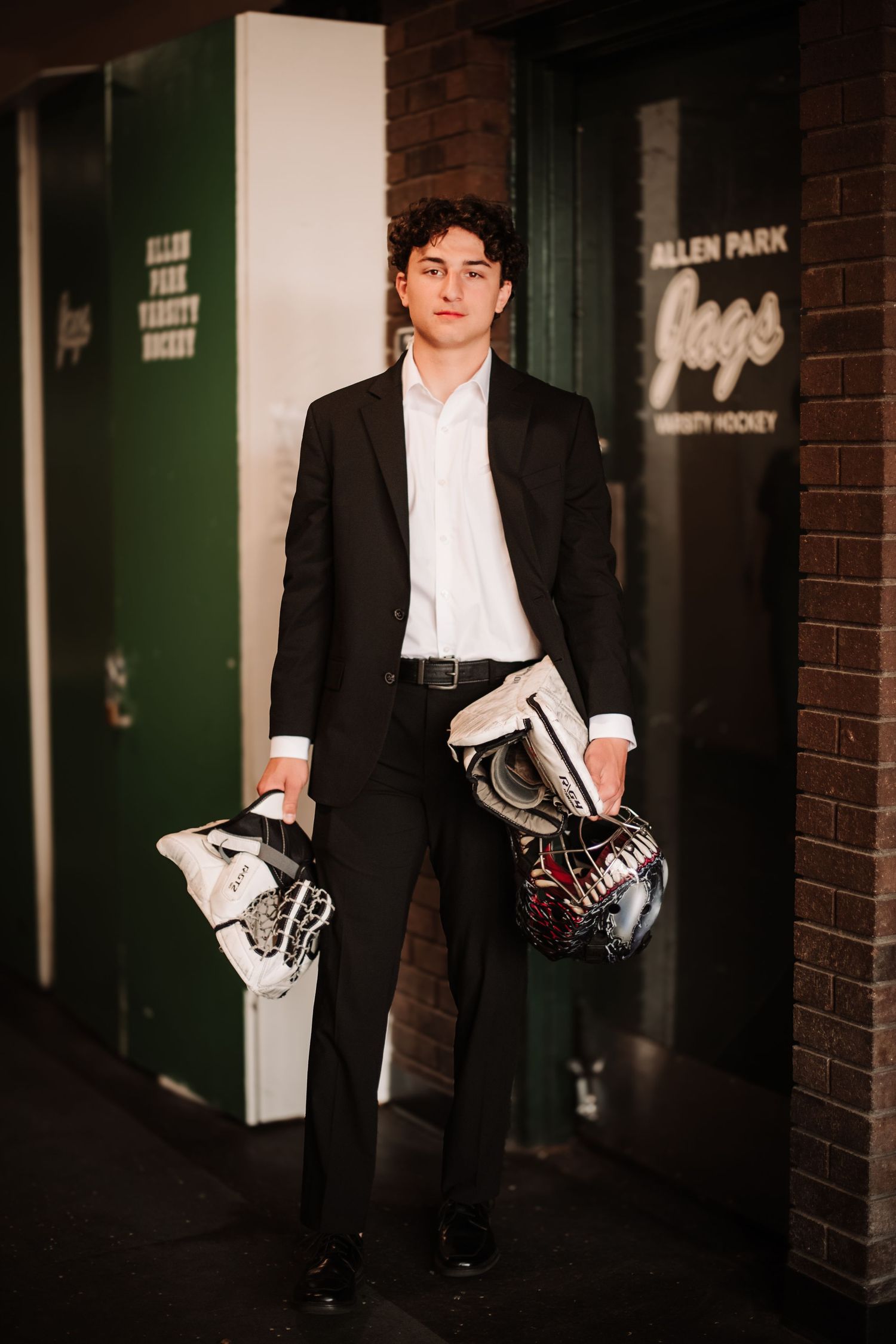 Young hockey player in black suit holding helmet and gloves outside Allen Park Varsity Hockey facility.