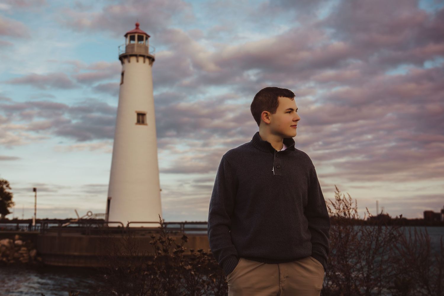 Young man standing near a white lighthouse at sunset with dramatic cloudy sky in the background.