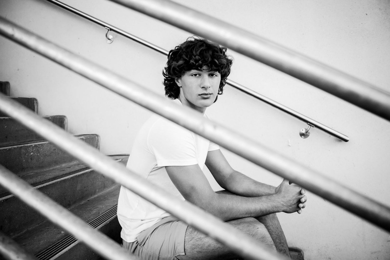 Young person with curly hair sitting on stairs in black and white photo wearing white t-shirt.