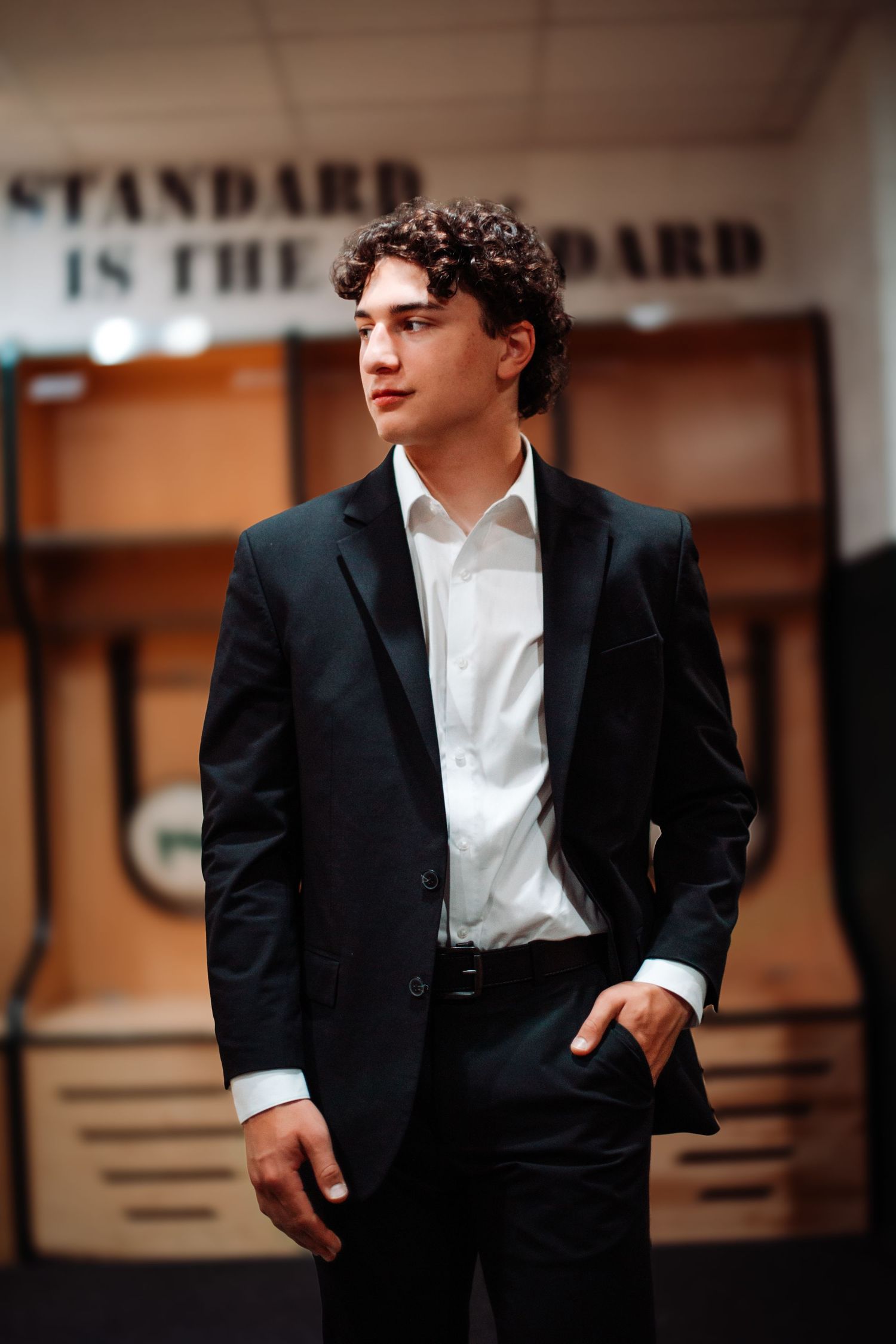 Young man with curly hair in a black suit and white shirt stands confidently in a modern gym setting.