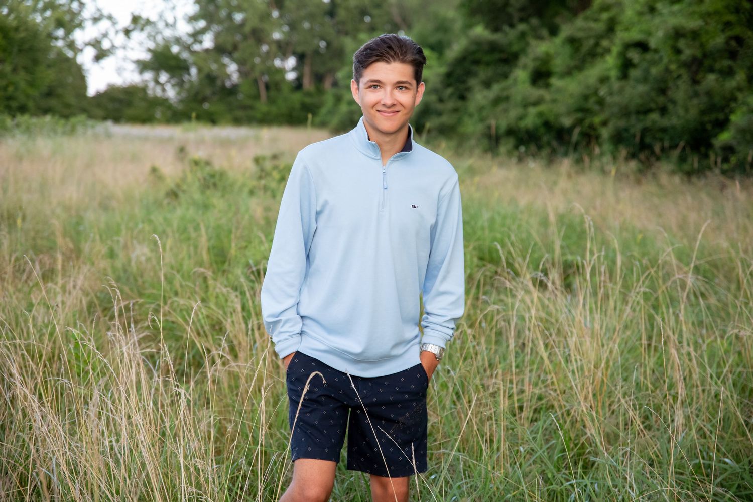Smiling teenage boy in light blue pullover and navy shorts standing in a lush green field outdoors.