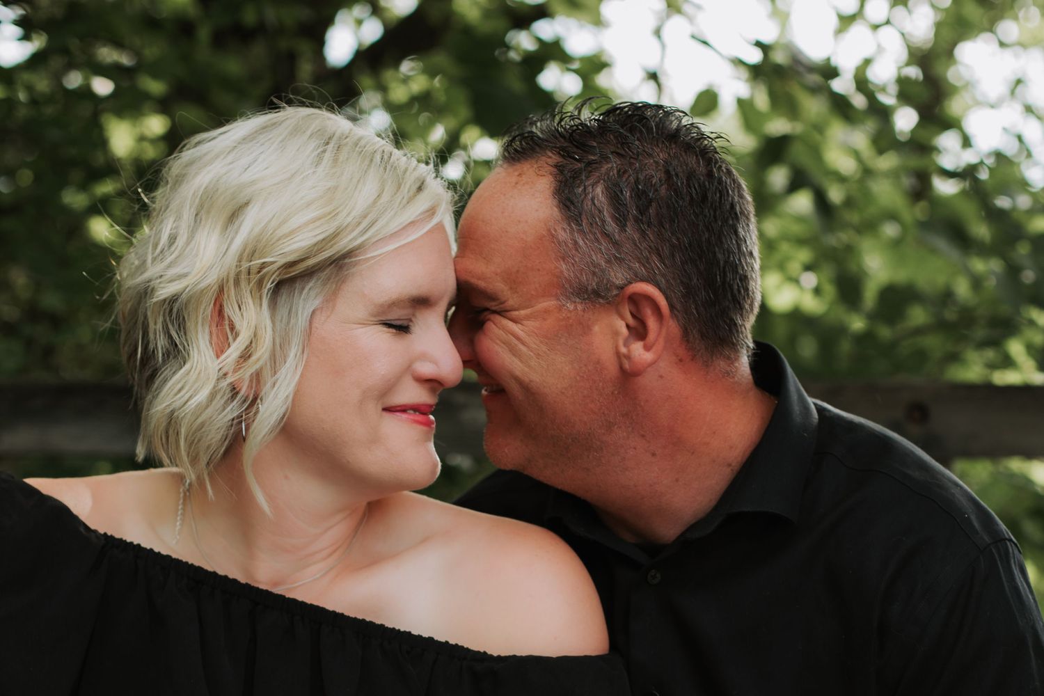 Couple sharing a tender moment close together outdoors with green trees behind them.