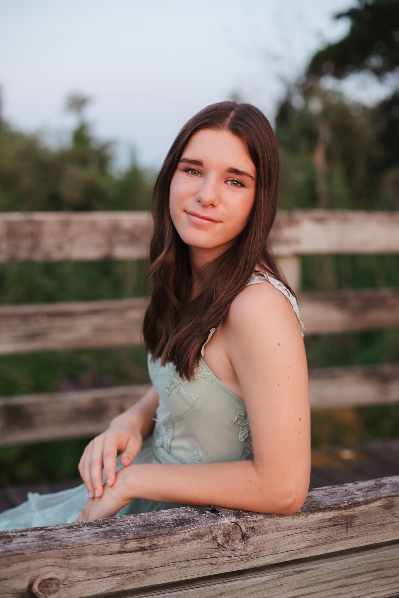 Young brunette teen girl in mint dress leaning on wooden fence outdoors at golden hour.