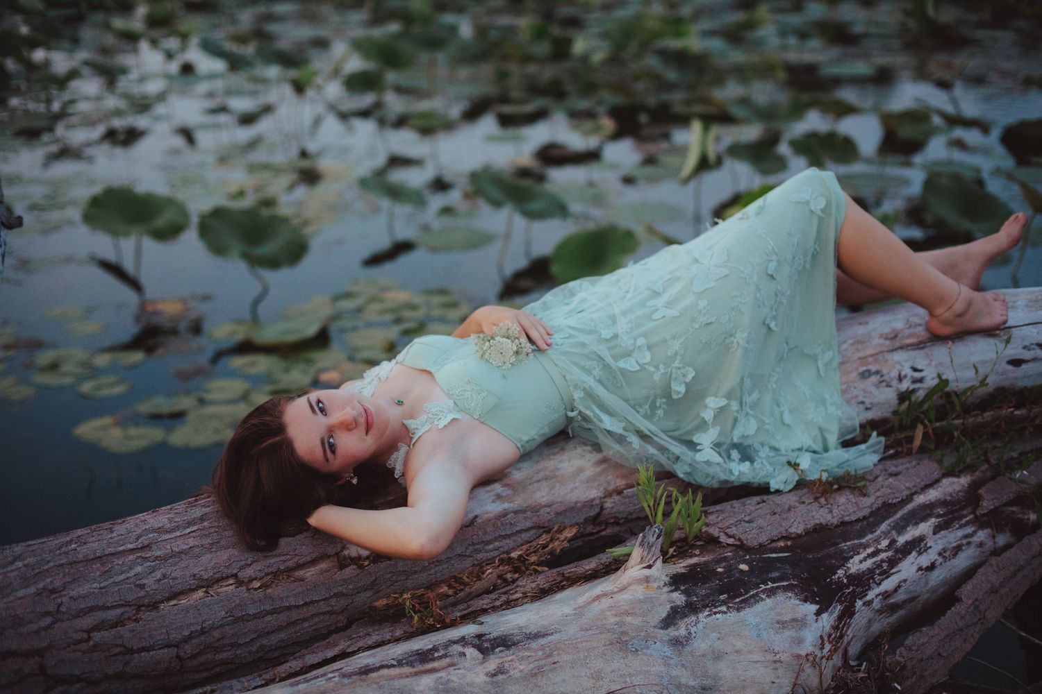 Young woman in mint lace dress lying on log surrounded by lily pads on serene pond.