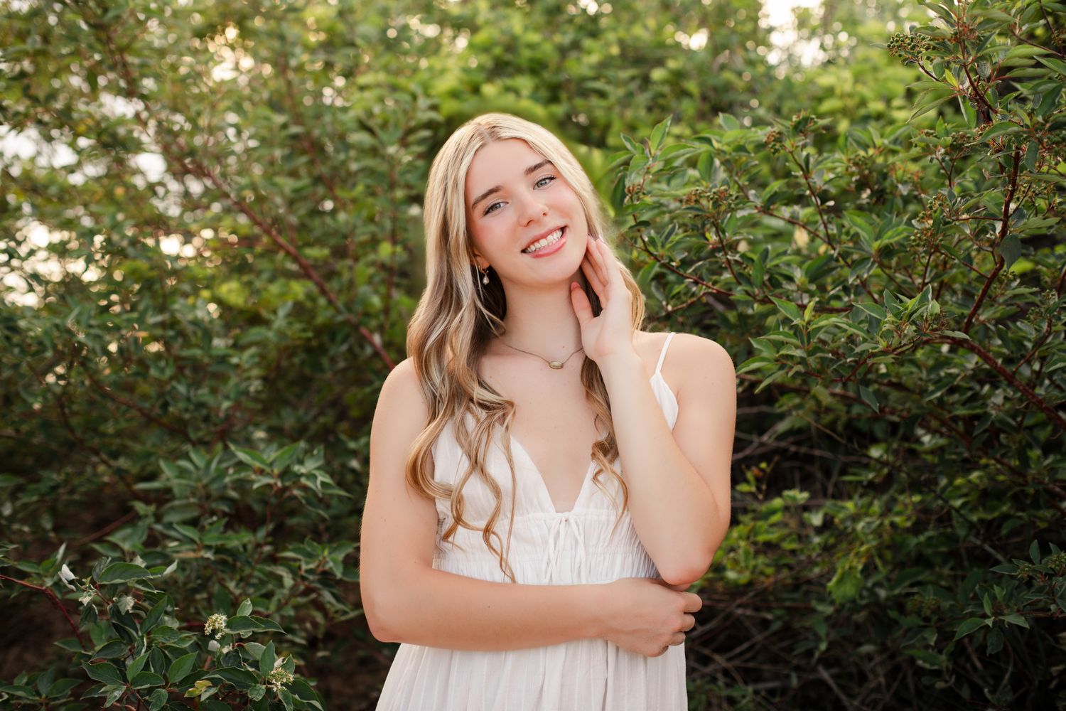 Smiling blonde woman in white dress posing outdoors among lush green bushes.