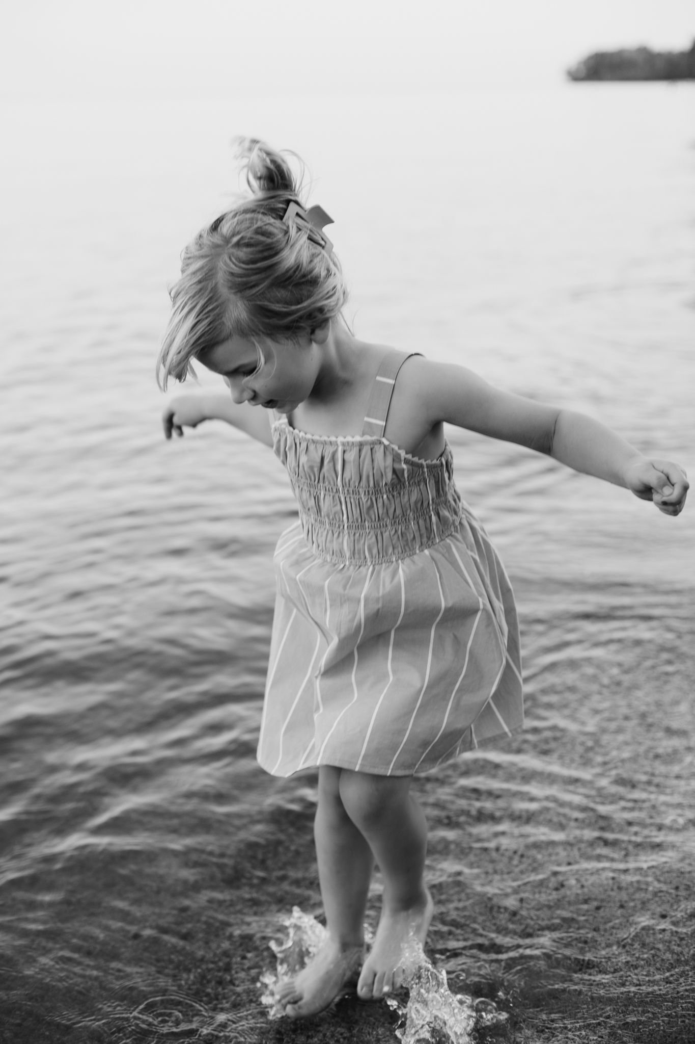 Black and white photo of a young girl in a striped dress splashing barefoot in shallow water near a lake shore.