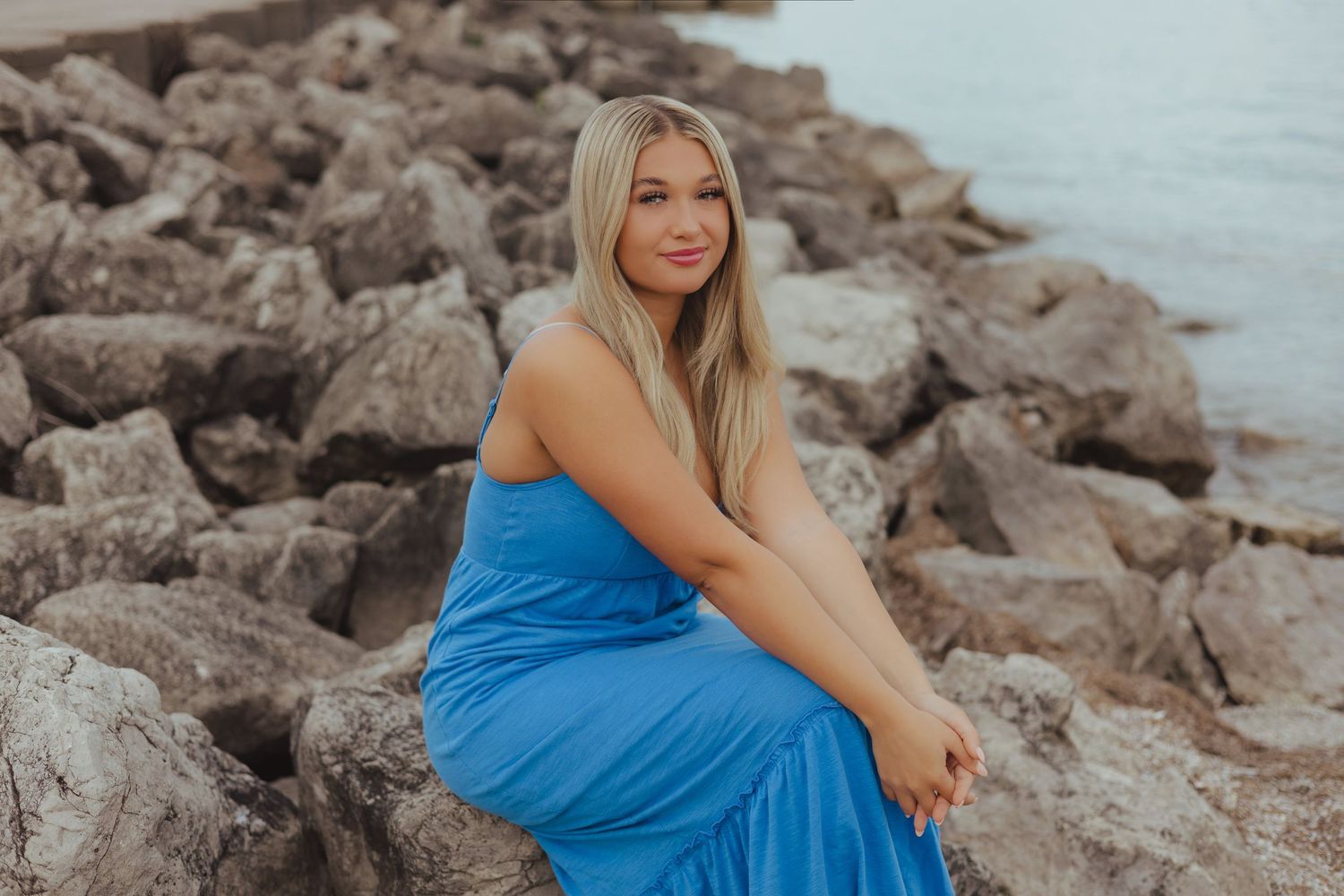 Young blonde woman in blue maxi dress posing on rocky shoreline near water.