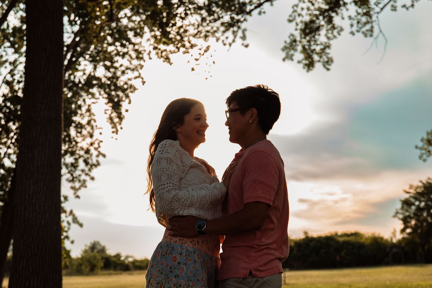 Couple embracing at golden hour sunset in a lush green park, sharing a romantic moment under a large tree.