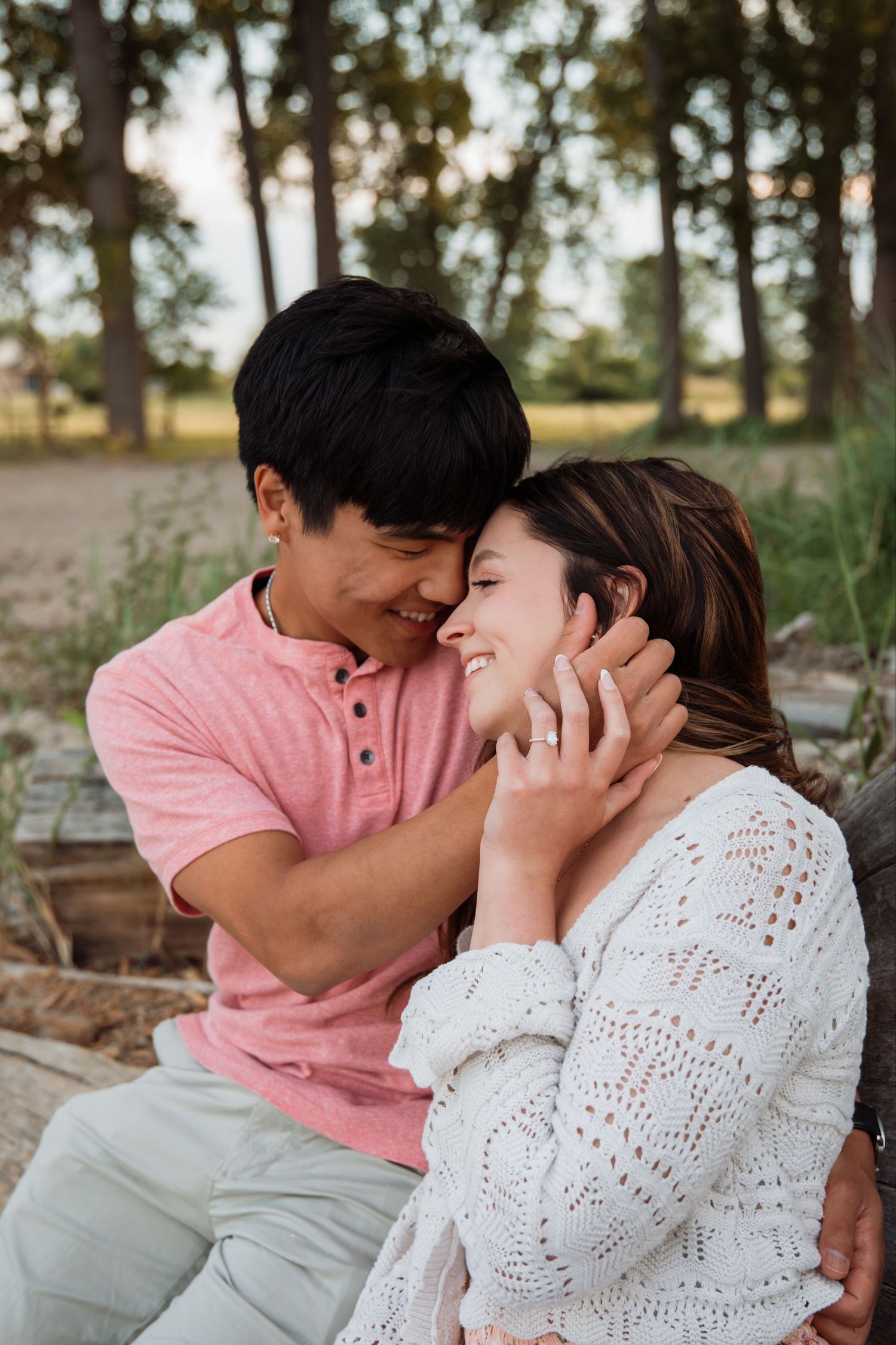 Smiling couple sharing a tender moment outdoors, man gently holding woman's face near sandy beach with trees in background.