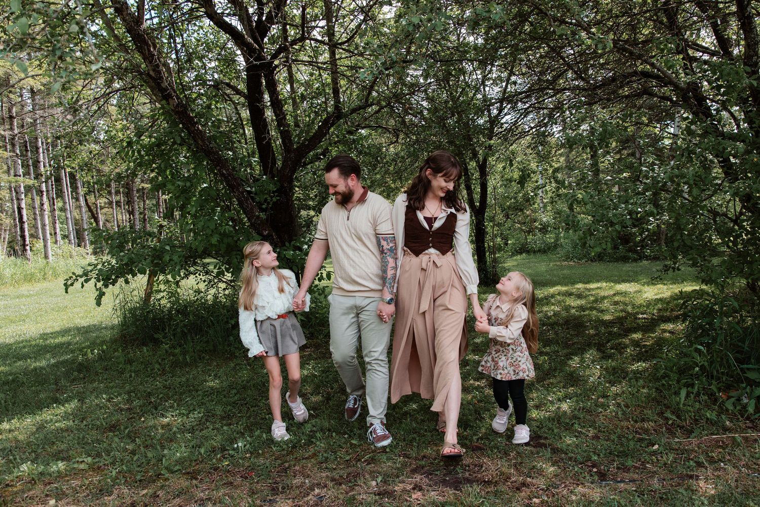 Family of four walking hand-in-hand through a lush green park, parents and two young daughters enjoying nature together.