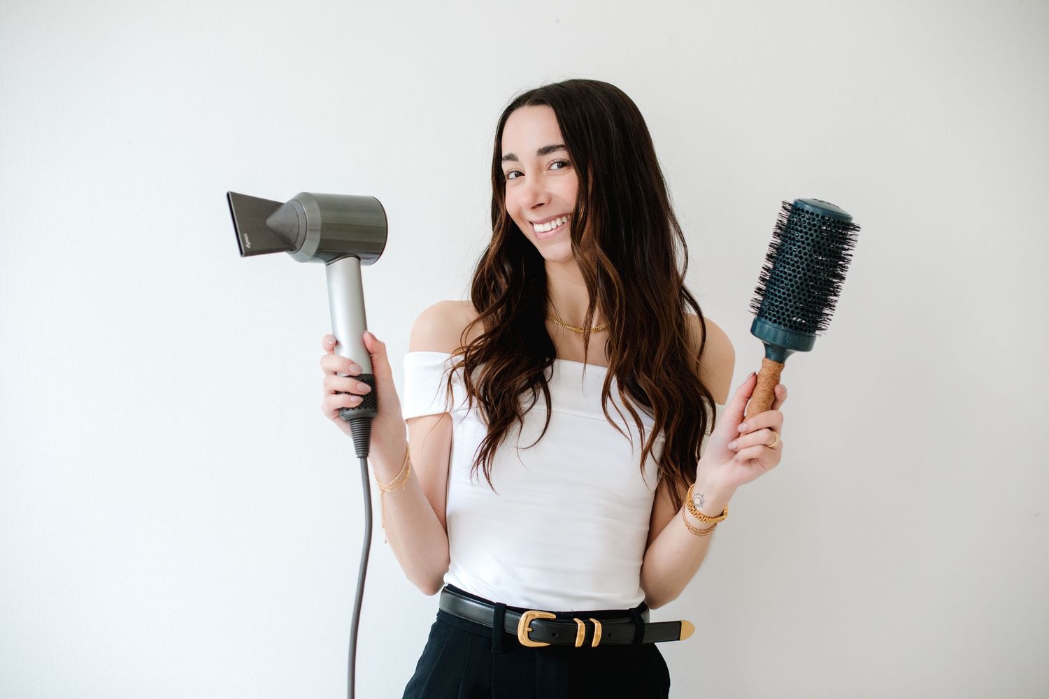 Smiling woman holding hair dryer and round brush wearing white off-shoulder top and gold belt.