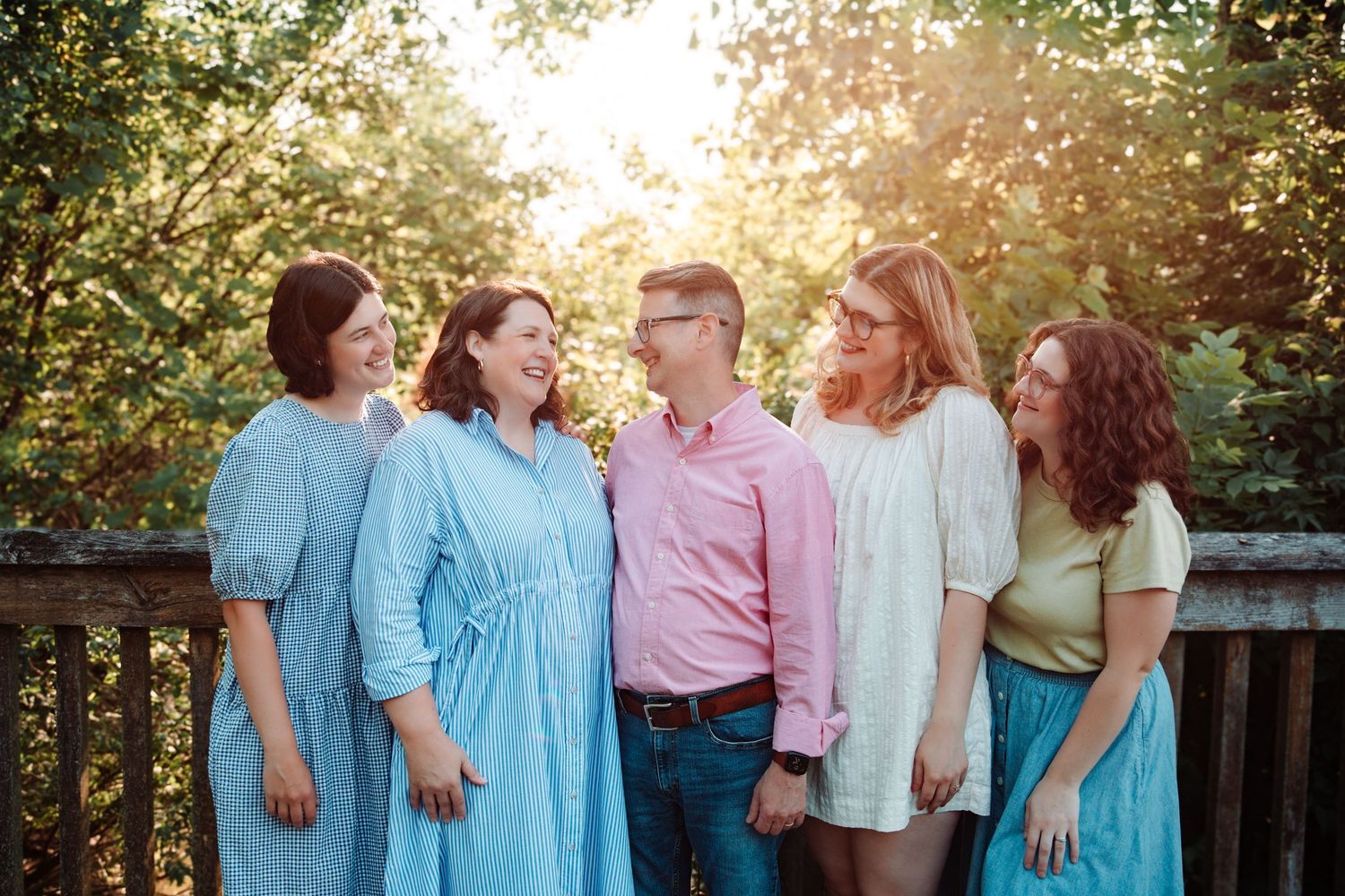 Family of five smiling and laughing together outdoors on a wooden deck surrounded by lush green trees.