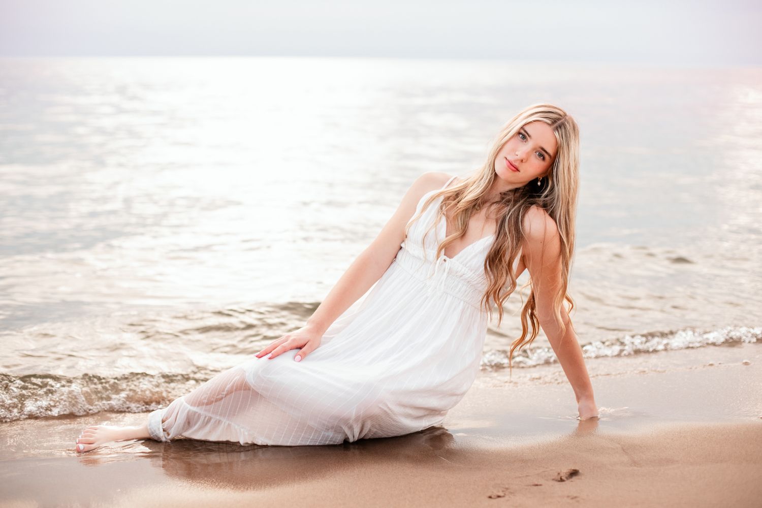 Young woman in a white dress sitting on a beach with gentle waves behind her.