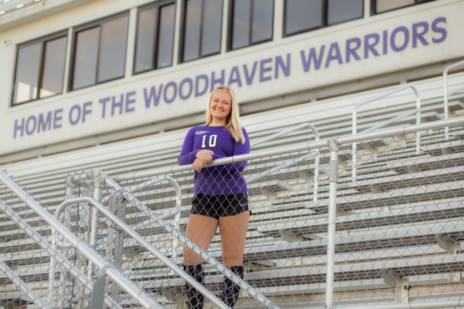 Female athlete in purple Woodhaven Warriors jersey number 10 posing on stadium bleachers.