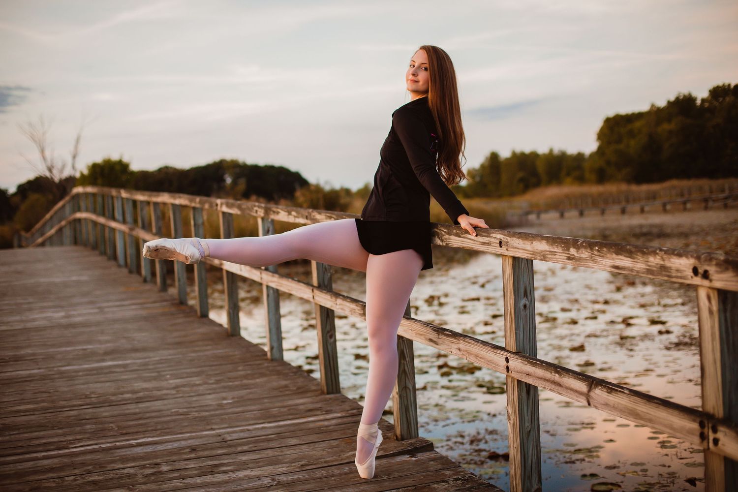 Ballerina in black outfit and pointe shoes posing at railing on scenic waterfront boardwalk.