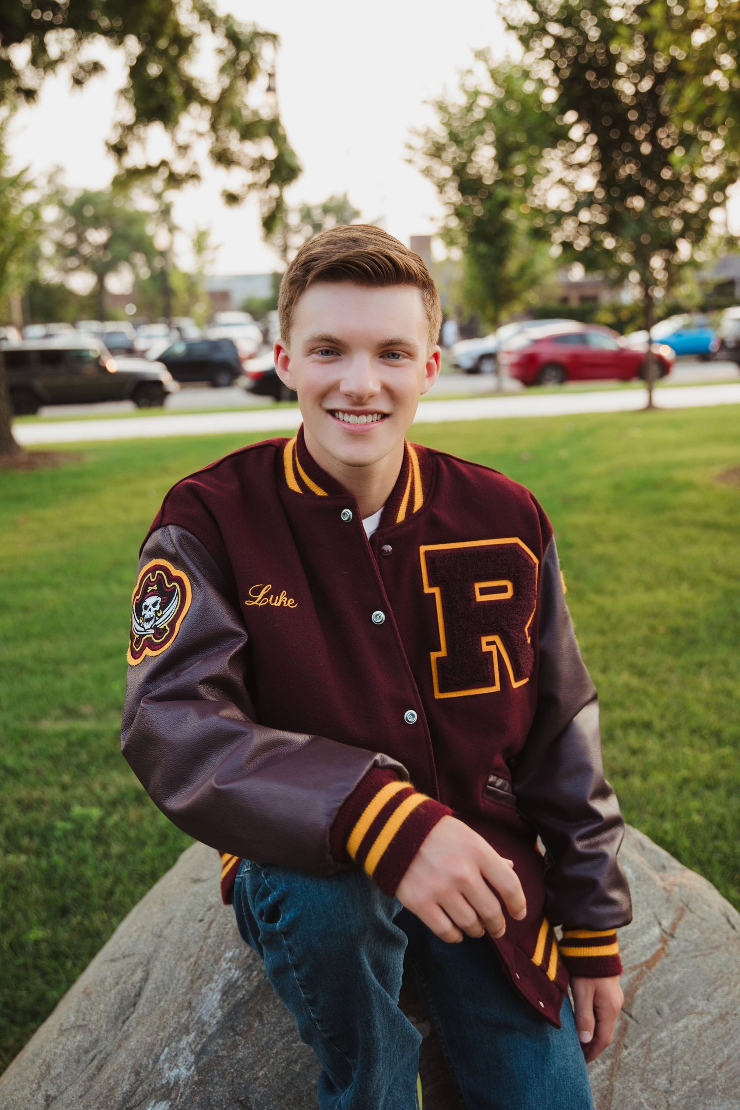 Smiling teen in maroon varsity letterman jacket sitting on rock in park for senior photos.