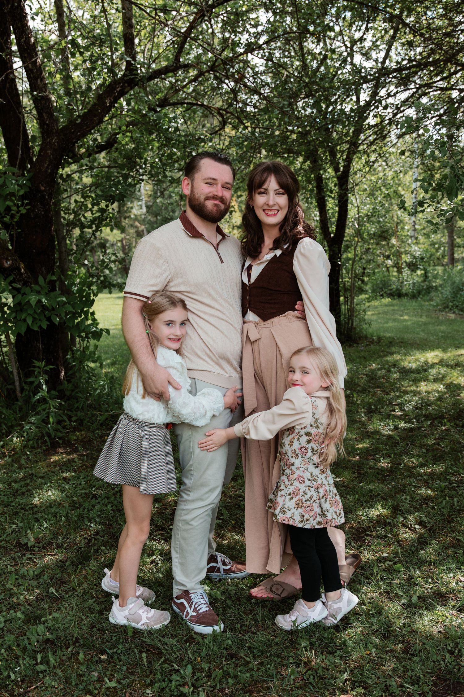 Happy family of four posing outdoors in a lush green garden, parents smiling with two young daughters hugging them.